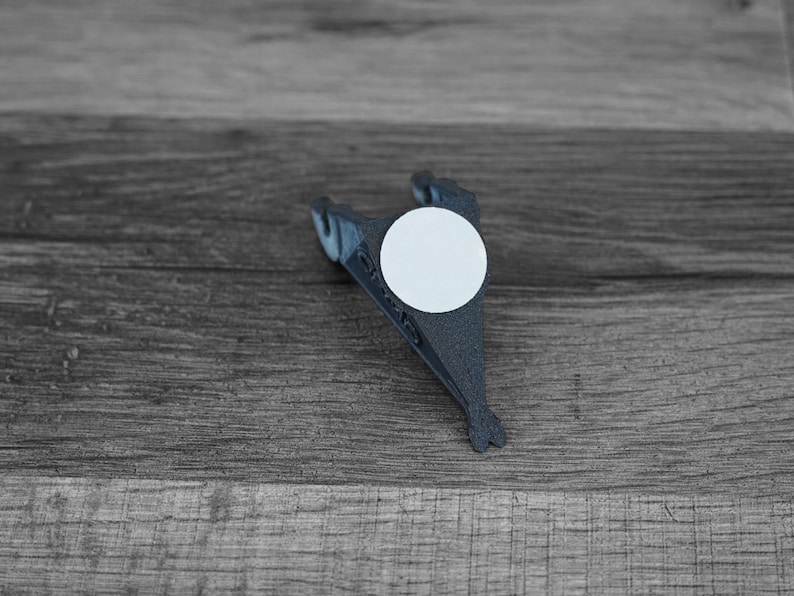 Puede incluir: Un objeto gris oscuro impreso en 3D con una pegatina circular blanca. El objeto tiene una forma &uacute;nica, posiblemente un clip o soporte, con un dise&ntilde;o en forma de aleta. El art&iacute;culo est&aacute; sobre un fondo de madera.