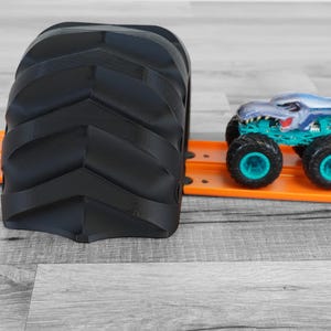 Peut inclure: Un obstacle de jouet en forme de pneu noir textur&eacute; &agrave; c&ocirc;t&eacute; d'une piste de jouet orange. Un monster truck bleu et argent&eacute; avec des roues turquoise est sur la piste. L'ensemble de jouets est sur une surface en bois grise.