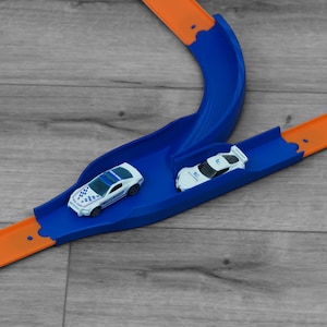 May include: A blue and orange toy race track with two white toy cars. One car has a police car design. The track has a curved blue section and straight orange sections. The cars are on the track.