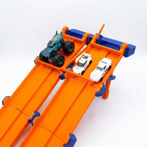 K&ouml;nnte beinhalten: Eine orangefarbene und blaue Spielzeug-Rennstrecke mit drei Spielzeugautos. Eines ist ein Monstertruck mit t&uuml;rkisblauem K&ouml;rper und schwarzen Reifen. Die anderen beiden sind wei&szlig;e Rennwagen.