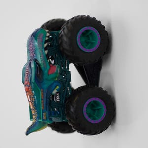 Puede incluir: Un cami&oacute;n monstruo de juguete con un dise&ntilde;o de dinosaurio turquesa y morado. El cami&oacute;n tiene neum&aacute;ticos negros grandes con detalles en verde y morado. La carrocer&iacute;a del cami&oacute;n es turquesa y morada con una cabeza de dinosaurio.