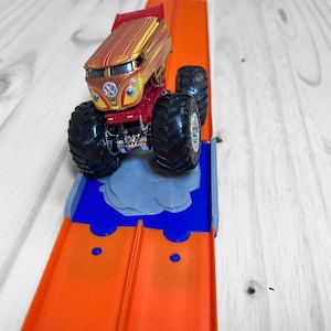 May include: A toy monster truck with a red, yellow and gold paint job is driving on an orange track. The truck has large black wheels and a white face with eyes and a mouth.
