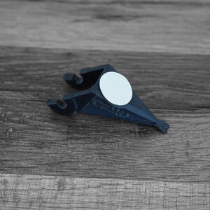 Puede incluir: Un objeto de pl&aacute;stico azul oscuro con una pegatina circular blanca. El objeto tiene una forma &uacute;nica con dos brazos curvos y un extremo puntiagudo. Est&aacute; colocado sobre un fondo de madera gris.