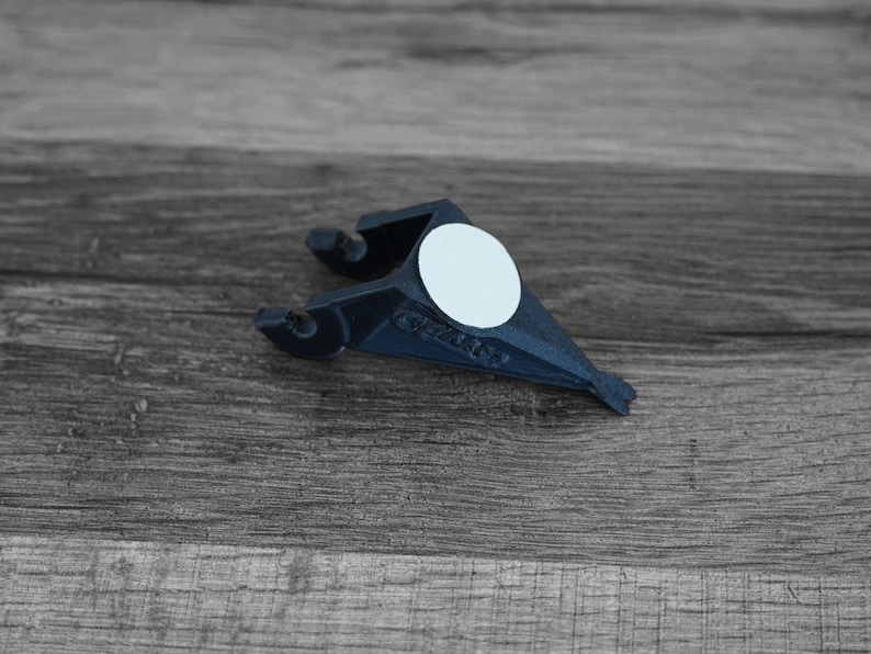 Puede incluir: Un objeto de pl&aacute;stico azul oscuro con una pegatina circular blanca. El objeto tiene una forma &uacute;nica con dos brazos curvos y un extremo puntiagudo. Est&aacute; colocado sobre un fondo de madera gris.