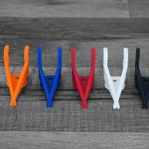 Puede incluir: Cinco clips de pl&aacute;stico en forma de V, de colores brillantes: naranja, azul, rojo, blanco y negro. Los clips est&aacute;n dispuestos en fila sobre una superficie de madera gris. Cada clip tiene un peque&ntilde;o agujero en el centro.