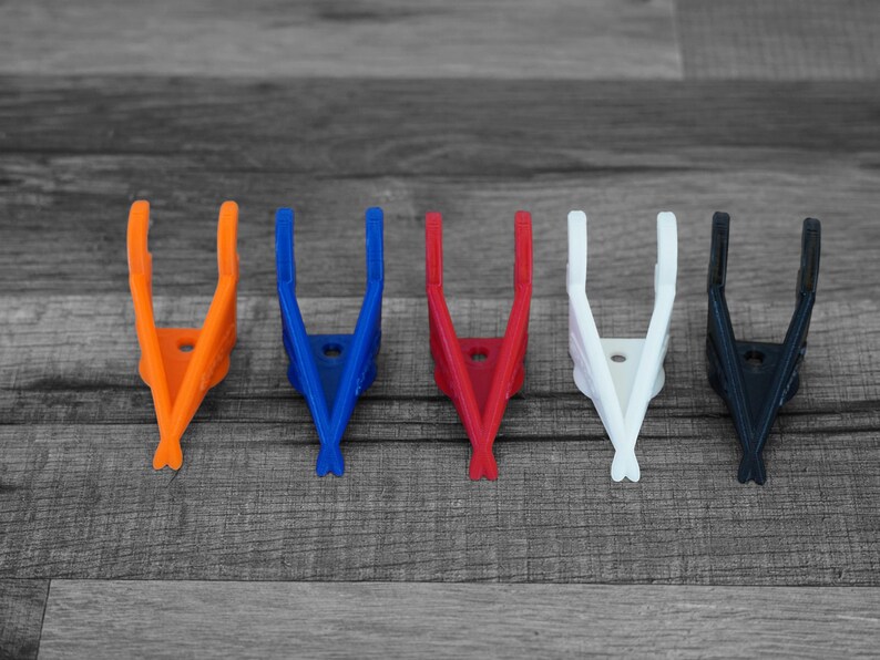 Puede incluir: Cinco clips de pl&aacute;stico en forma de V, de colores brillantes: naranja, azul, rojo, blanco y negro. Los clips est&aacute;n dispuestos en fila sobre una superficie de madera gris. Cada clip tiene un peque&ntilde;o agujero en el centro.