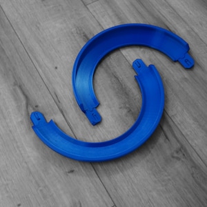 Puede incluir: Dos piezas de plástico azul brillante, curvadas, posiblemente partes de un juguete o una estructura más grande, están dispuestas sobre una superficie de madera gris. Las piezas tienen forma semicircular con pequeñas pestañas en los extremos.