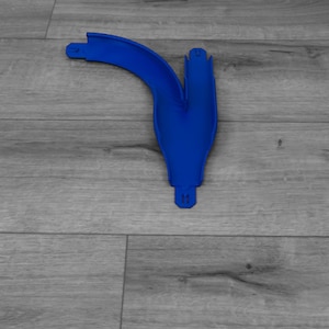 Puede incluir: Una pieza de pista de juguete de plástico azul brillante, con forma de Y curvada. La pista está colocada sobre una superficie de madera gris. La pieza tiene una sección curva y una sección dividida, con pequeños puntos de conexión en los extremos.
