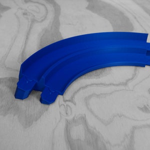 Puede incluir: Dos piezas de pista de plástico azul para una pista de carreras de coches de juguete. Las piezas son curvas y se conectan para formar una pista continua.