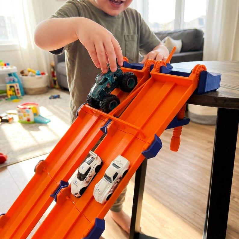K&ouml;nnte beinhalten: Eine orangefarbene und blaue Spielzeug-Rennstrecke mit drei Spielzeugautos. Die Strecke hat zwei Fahrspuren und ist nach oben abgewinkelt. Ein Kind spielt mit den Spielzeugautos. Einer ist ein Monstertruck und die anderen beiden sind wei&szlig;e Rennwagen.