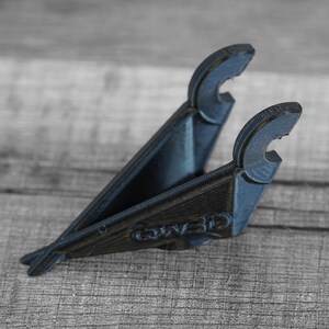 Puede incluir: Un soporte para tel&eacute;fono impreso en 3D de color azul oscuro. El soporte tiene forma triangular con dos brazos curvos en la parte superior para sujetar un tel&eacute;fono. La base tiene el texto "QW3D" impreso. El soporte est&aacute; sobre una superficie de madera.