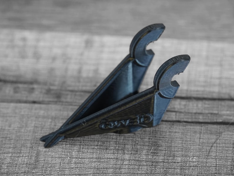 Puede incluir: Un soporte para tel&eacute;fono impreso en 3D de color azul oscuro. El soporte tiene forma triangular con dos brazos curvos en la parte superior para sujetar un tel&eacute;fono. La base tiene el texto "QW3D" impreso. El soporte est&aacute; sobre una superficie de madera.