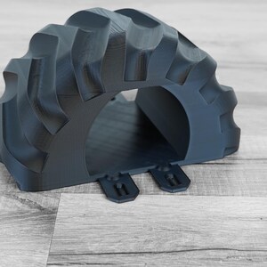 Peut inclure: Un passage en forme d'arche de pneu de tracteur imprim&eacute; en 3D, de couleur gris fonc&eacute;. La bande de roulement du pneu est visible sur le dessus et les c&ocirc;t&eacute;s, avec une ouverture arrondie au centre. Deux languettes rectangulaires sont &agrave; la base.