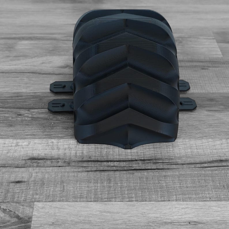 Peut inclure: Un objet noir incurv&eacute; et textur&eacute; avec une s&eacute;rie de cr&ecirc;tes en forme de chevrons. Deux petites languettes rectangulaires s'&eacute;tendent des c&ocirc;t&eacute;s. L'objet est pos&eacute; sur une surface en bois grise.