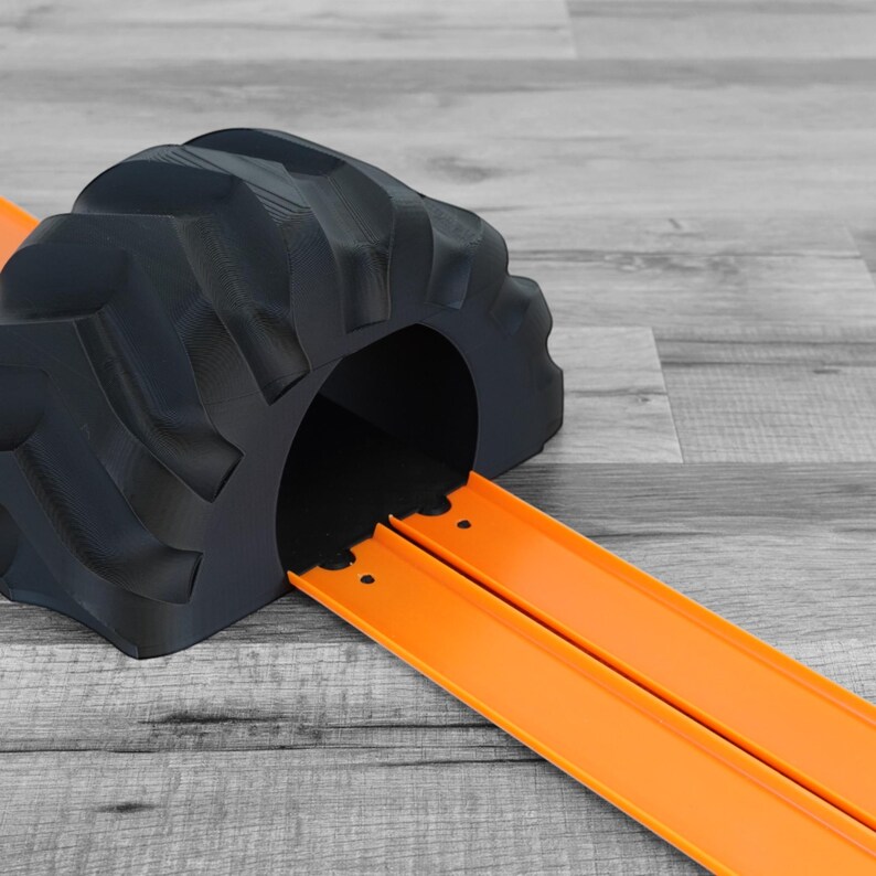 Peut inclure: Un tunnel de jouet noir imprim&eacute; en 3D en forme de pneu, avec une piste de course orange s'&eacute;tendant de l'ouverture du tunnel. La piste est con&ccedil;ue pour les voitures jouets. Le tunnel est sur une surface en bois grise.