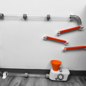 May include: A clear plastic tube and orange and white plastic track toy set against a white wall. The set includes a funnel, a white base with a gray handle, and several orange tubes. The toy is designed to be mounted on a wall.