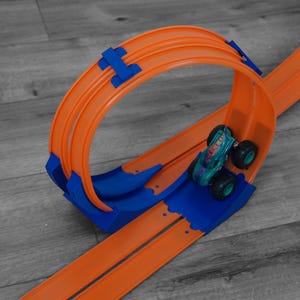 Könnte beinhalten: Eine orangefarbene und blaue Spielzeug-Rennstrecke mit Looping-Design. Ein tealfarbener Monstertruck mit schwarzen Rädern befindet sich auf der Strecke. Die Rennstrecke ist aus Kunststoff und liegt auf einer grauen Holzoberfläche.