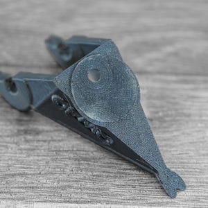 Puede incluir: Objeto gris oscuro, impreso en 3D, con una forma &uacute;nica, que presenta un elemento circular y una cola en forma de aleta. El objeto tiene una superficie texturizada y parece ser un art&iacute;culo decorativo o funcional.