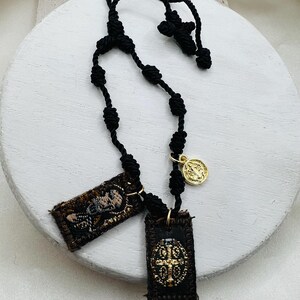 May include: Black knotted rosary with two rectangular pendants and a gold-colored medallion. The pendants feature embroidered designs in brown and gold. The rosary is displayed on a white, round surface.