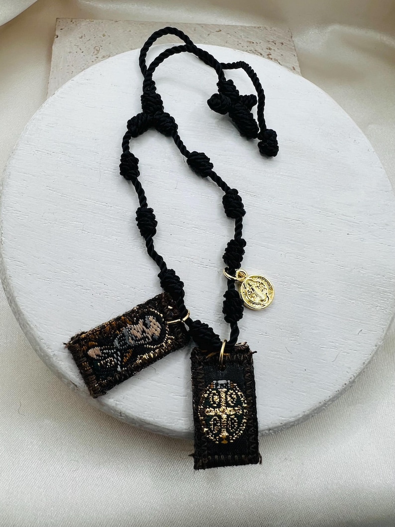 May include: Black knotted rosary with two rectangular pendants and a gold-colored medallion. The pendants feature embroidered designs in brown and gold. The rosary is displayed on a white, round surface.