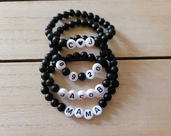 May include: Three black beaded bracelets with white letter beads spelling out "CJ", "8920", and "JACOB" with a heart bead between the "C" and "J" on the top bracelet. The bottom bracelet also has the word "MAMA" spelled out.