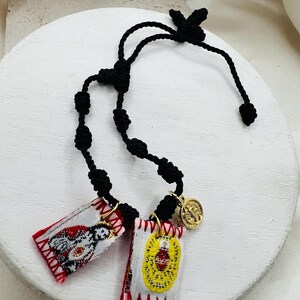 May include: A black cord rosary with two small rectangular pendants and a gold-colored medallion. The pendants feature embroidered designs in red, white, and yellow. The medallion has an image of a saint.