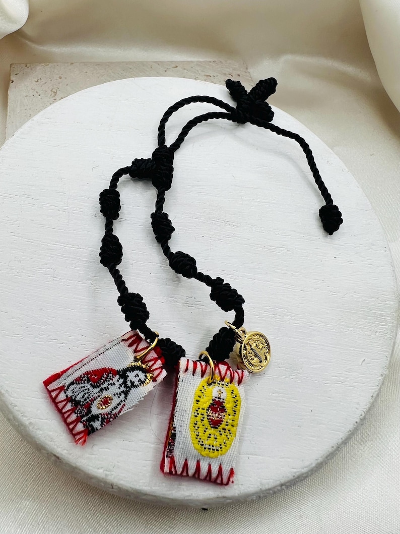 May include: A black cord rosary with two small rectangular pendants and a gold-colored medallion. The pendants feature embroidered designs in red, white, and yellow. The medallion has an image of a saint.