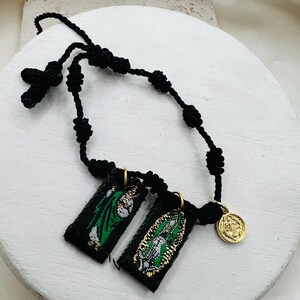 May include: A black corded rosary with a cross and knotted beads. Two small black rectangular fabric pendants with green and gold designs hang from the rosary. A gold-colored coin charm is also attached.