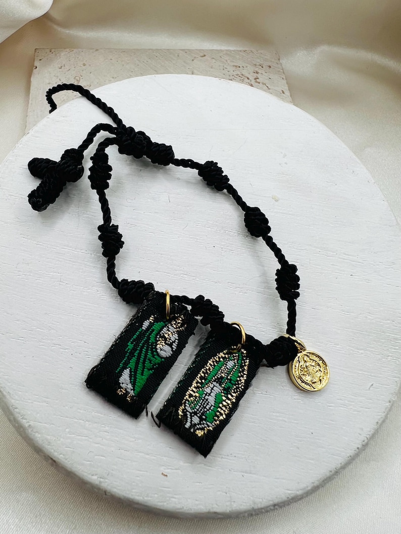 May include: A black corded rosary with a cross and knotted beads. Two small black rectangular fabric pendants with green and gold designs hang from the rosary. A gold-colored coin charm is also attached.