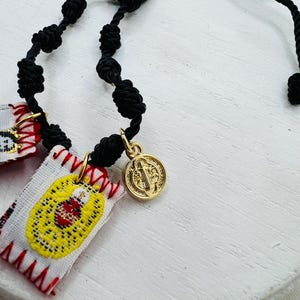 May include: A black knotted cord rosary with a gold-colored medallion and small embroidered patches. The patches are white with yellow and red embroidered designs. The medallion features a religious figure.