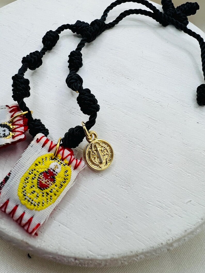 May include: A black knotted cord rosary with a gold-colored medallion and small embroidered patches. The patches are white with yellow and red embroidered designs. The medallion features a religious figure.