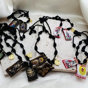 May include: Several black rosaries with small, rectangular embroidered pendants featuring religious imagery and gold-colored coin charms. The pendants display various saints and religious scenes. The rosaries are arranged on a light-colored surface.