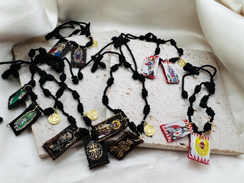 May include: Several black rosaries with small, rectangular embroidered pendants featuring religious imagery and gold-colored coin charms. The pendants display various saints and religious scenes. The rosaries are arranged on a light-colored surface.
