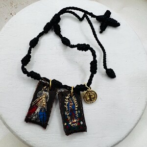 May include: A black cord rosary bracelet featuring two rectangular fabric pendants with religious imagery and a gold-colored medallion. The pendants depict detailed illustrations with gold, red, blue, and white accents. The bracelet is displayed on a white surface.