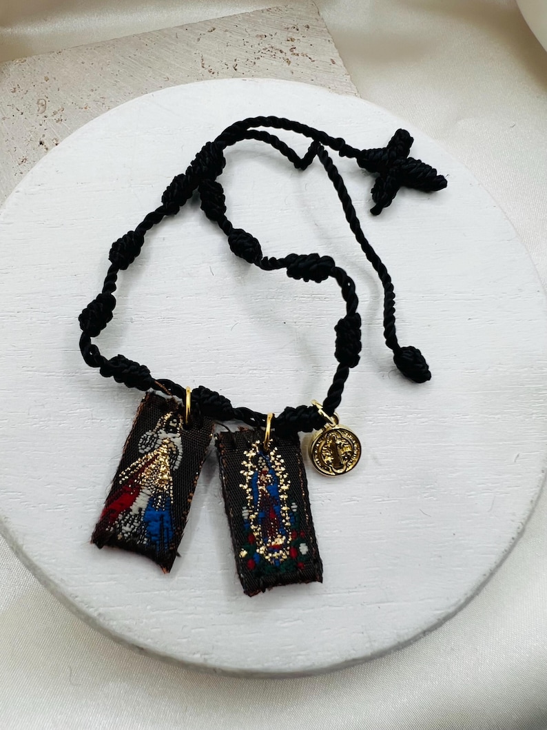 May include: A black cord rosary bracelet featuring two rectangular fabric pendants with religious imagery and a gold-colored medallion. The pendants depict detailed illustrations with gold, red, blue, and white accents. The bracelet is displayed on a white surface.