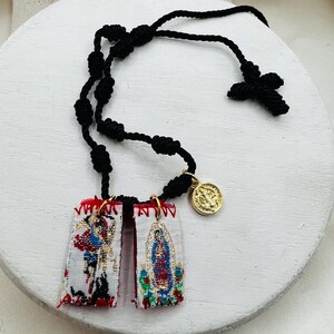 May include: A black knotted rosary with a cross and two rectangular embroidered pendants. The pendants feature colorful religious imagery and are connected to a gold-colored medallion. The rosary is displayed on a white surface.