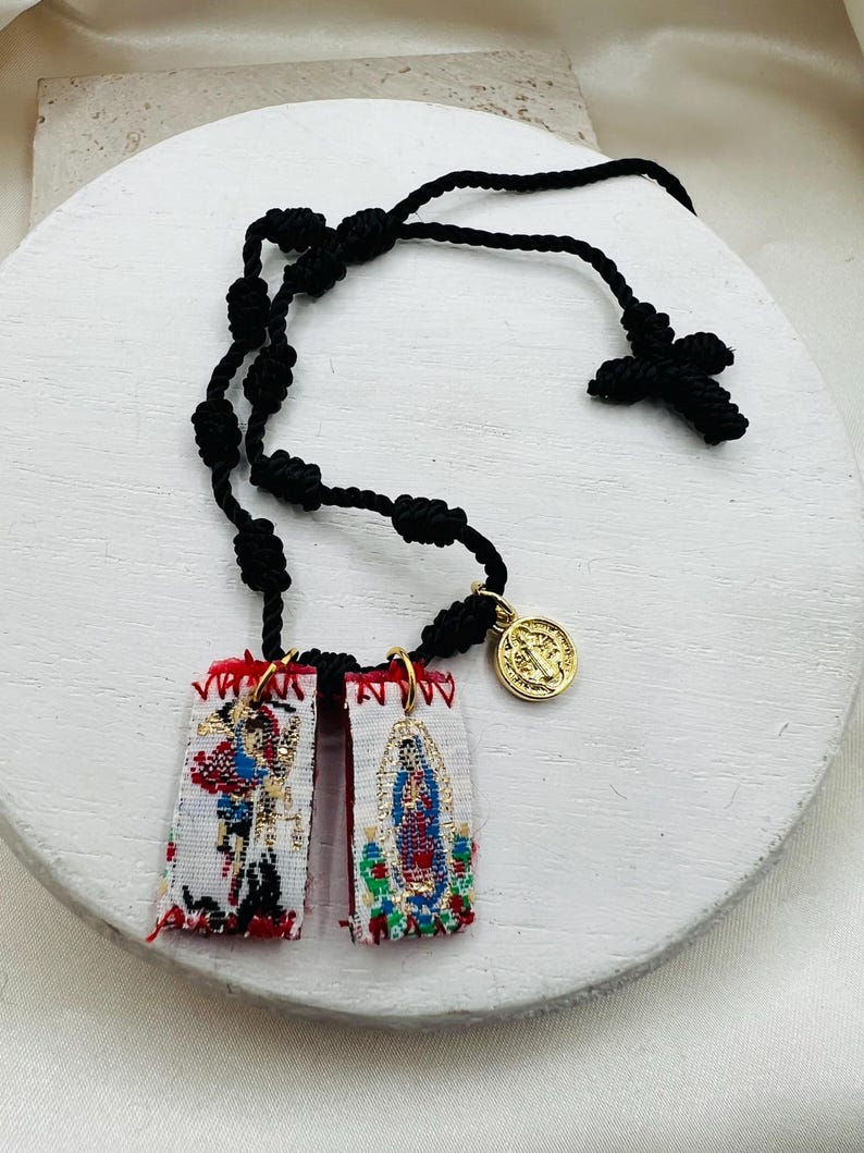 May include: A black knotted rosary with a cross and two rectangular embroidered pendants. The pendants feature colorful religious imagery and are connected to a gold-colored medallion. The rosary is displayed on a white surface.