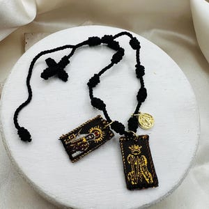 Può includere: Un rosario nero con una croce e grani annodati. Due pendenti rettangolari ricamati con immagini religiose sono appesi al rosario, insieme a una piccola moneta color oro.