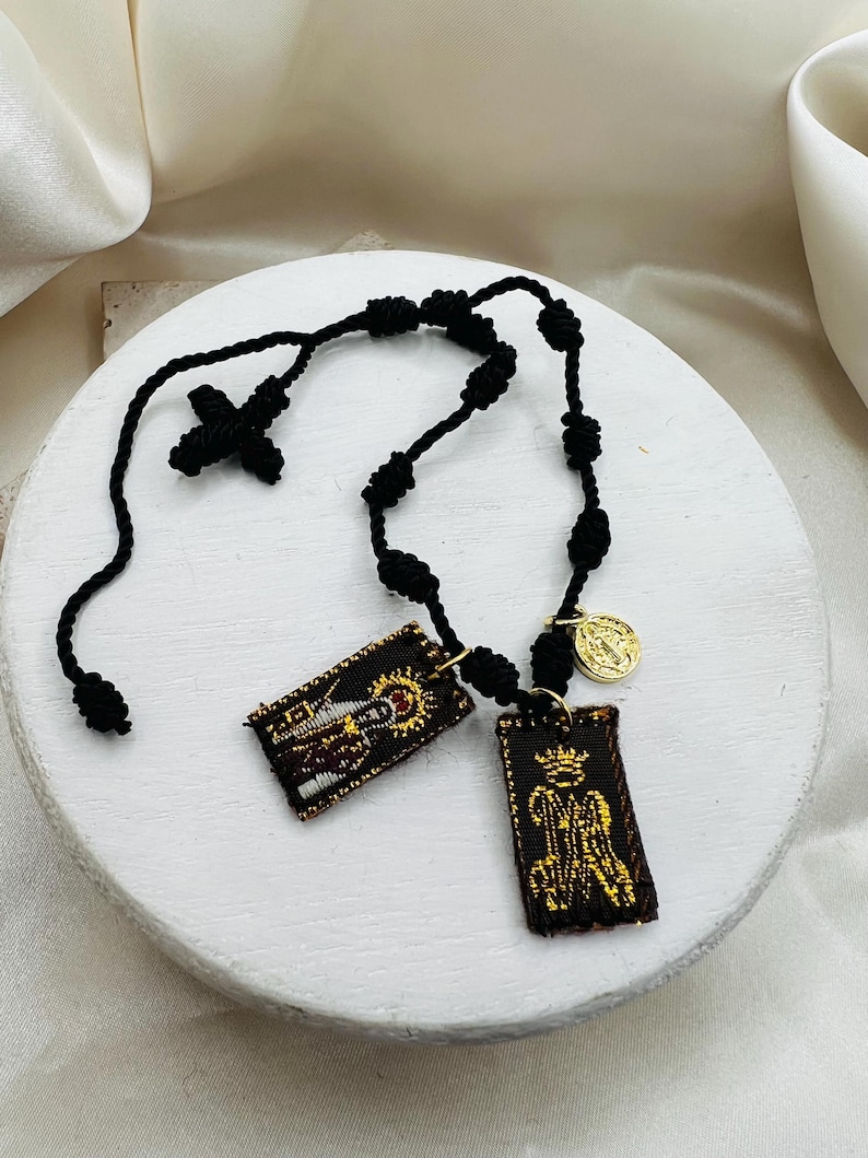 May include: A black rosary with a cross and knotted beads. Two rectangular embroidered pendants with religious imagery hang from the rosary, along with a small gold-colored coin.