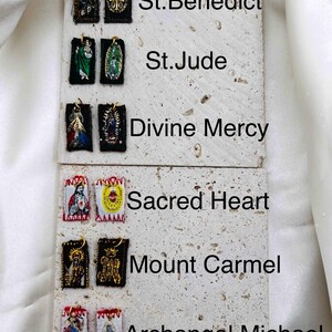 May include: A collection of religious pendants featuring various saints, including St. Benedict, St. Jude, and Archangel Michael. Each pendant is rectangular, with detailed embroidered images and gold-colored accents. The pendants are displayed on a textured, light-colored surface.