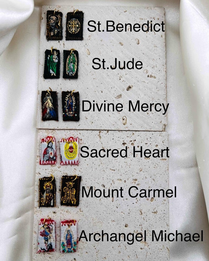 May include: A collection of religious pendants featuring various saints, including St. Benedict, St. Jude, and Archangel Michael. Each pendant is rectangular, with detailed embroidered images and gold-colored accents. The pendants are displayed on a textured, light-colored surface.