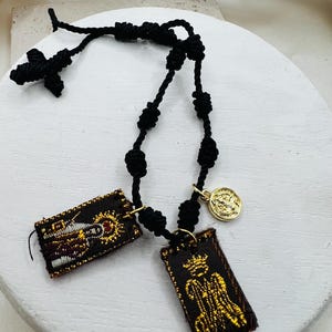 May include: A black knotted cord rosary with a small black cross and two rectangular fabric pendants. The pendants feature gold embroidery on a dark background. A small gold coin charm is also attached to the rosary.