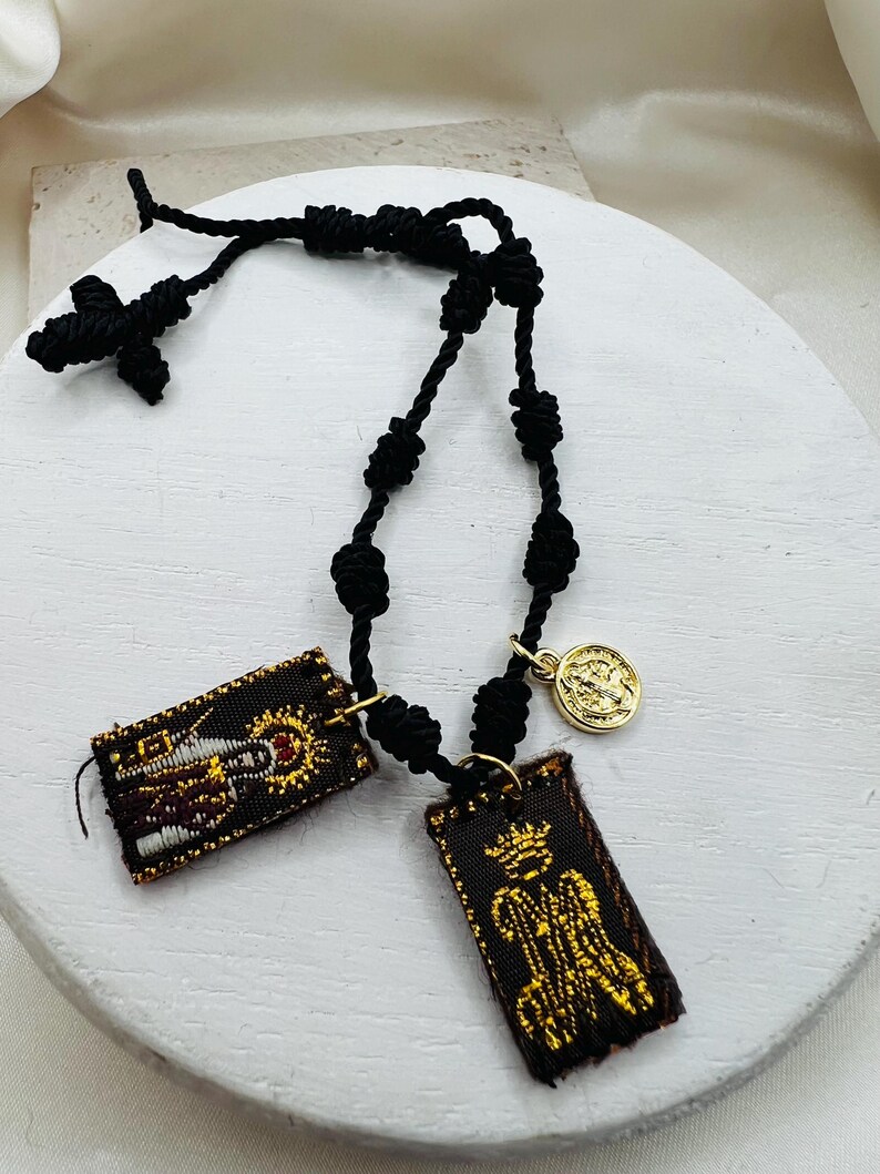 May include: A black knotted cord rosary with a small black cross and two rectangular fabric pendants. The pendants feature gold embroidery on a dark background. A small gold coin charm is also attached to the rosary.