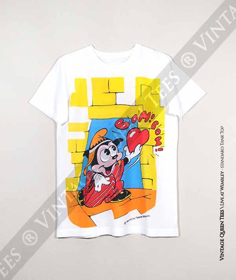 May include: White t-shirt featuring a cartoon image of Mickey Mouse wearing a red and white striped outfit and holding a red heart. The text "Boom! Boom!" is above Mickey Mouse. The background is yellow, blue, and orange.