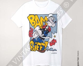 May include: A white short-sleeve t-shirt featuring a colorful comic book style graphic of Popeye the Sailor. The print includes the bold yellow text BAM, POW, and BIFF, along with blue dot patterns, red accents, and the character in action.
