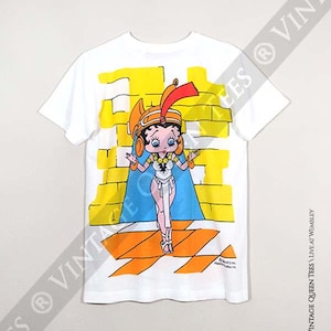 May include: White t-shirt featuring a cartoon image of Betty Boop dressed as an Egyptian queen. The image is printed on the back of the shirt and has a yellow and orange background.