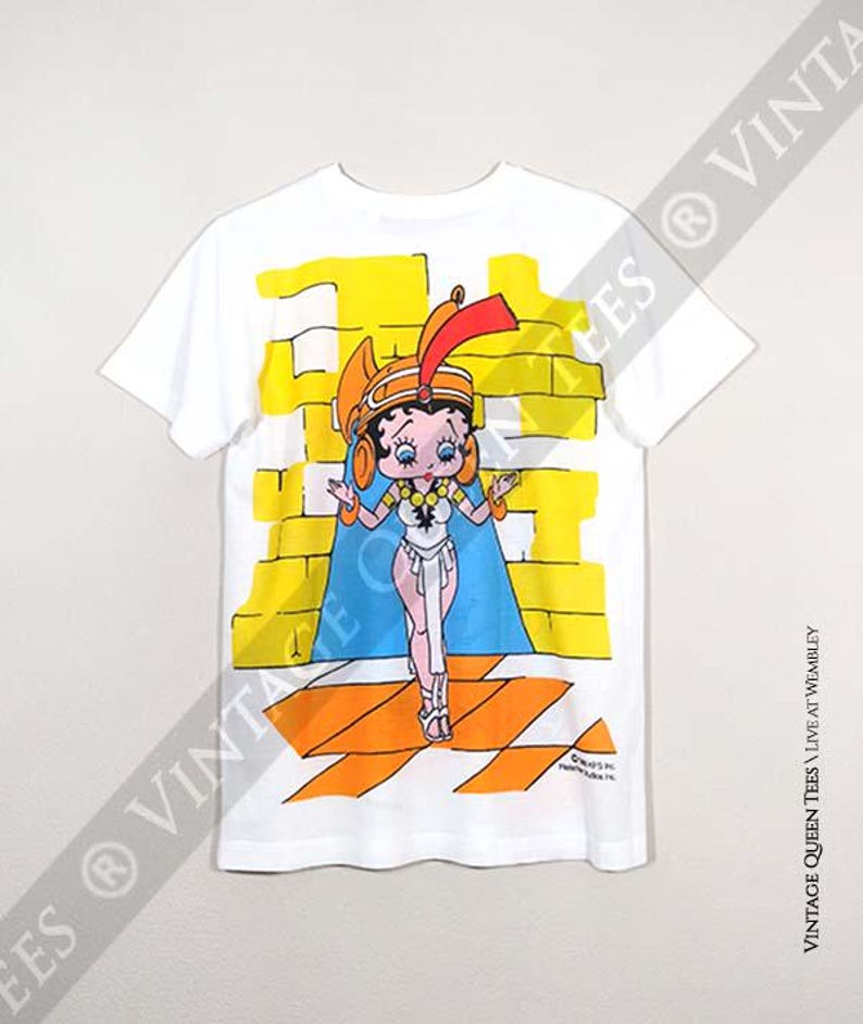 May include: White t-shirt featuring a cartoon image of Betty Boop dressed as an Egyptian queen. The image is printed on the back of the shirt and has a yellow and orange background.