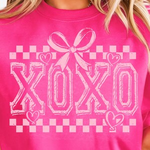 Puede incluir: Una sudadera rosa con un patr&oacute;n de cuadros blancos y la palabra "XOXO" en una fuente rosa, delineada. Un lazo rosa con un coraz&oacute;n a cada lado est&aacute; por encima de la palabra.