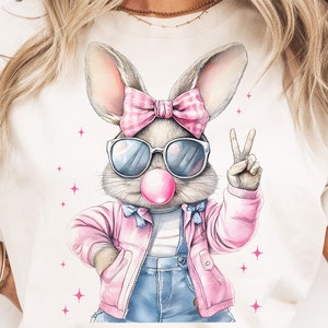 May include: A white t-shirt with a graphic of a cartoon bunny wearing pink sunglasses, a pink jacket, blue overalls, and a pink bow. The bunny is holding up two fingers in a peace sign and has a pink bubble gum in its mouth.