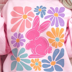May include: Pink sweatshirt with a graphic design of a pink bunny rabbit surrounded by colorful flowers. The flowers are in a retro style with a faded, vintage look.